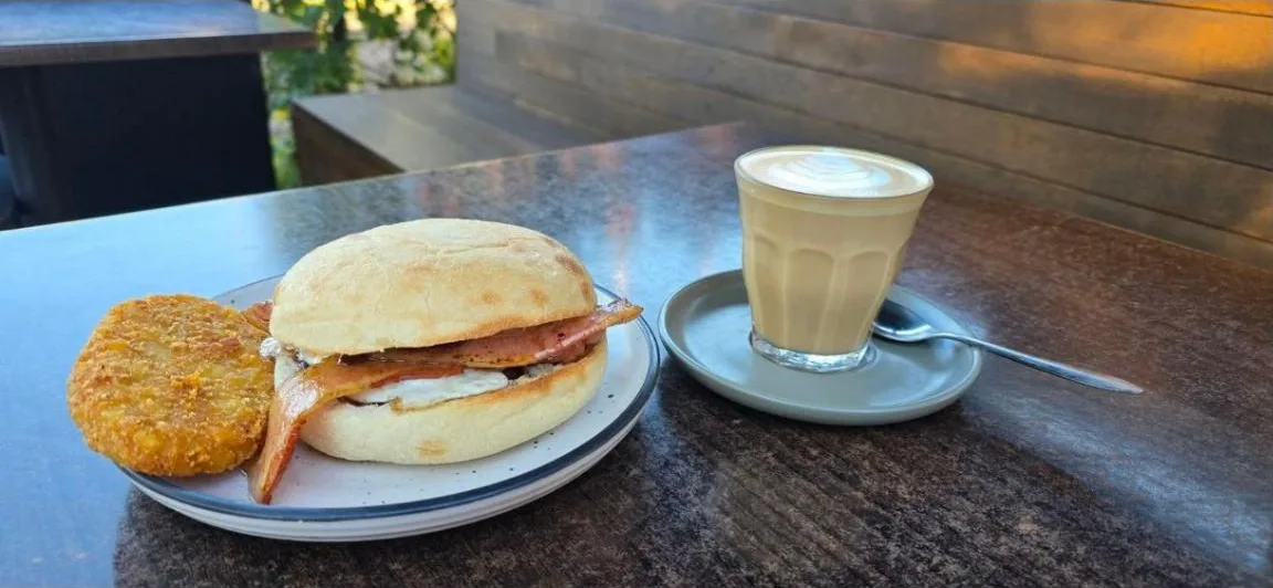 A bacon and egg roll with a hash brown and a latte